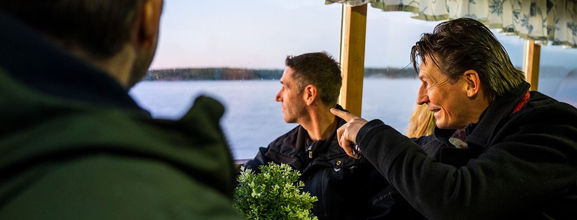 Tre män sitter inne ombord på en skärgårdsbåt. En av männen pekar ut mot skärgården.