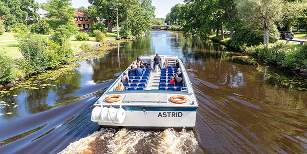 Båten M/F Astrid i lugnt vatten. I båten sitter flera personer. Föraren kör båten stående.