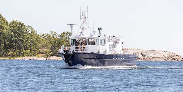 Båten M/F Tuva. Det är mulet väder och i aktern står flera personer och samtalar.