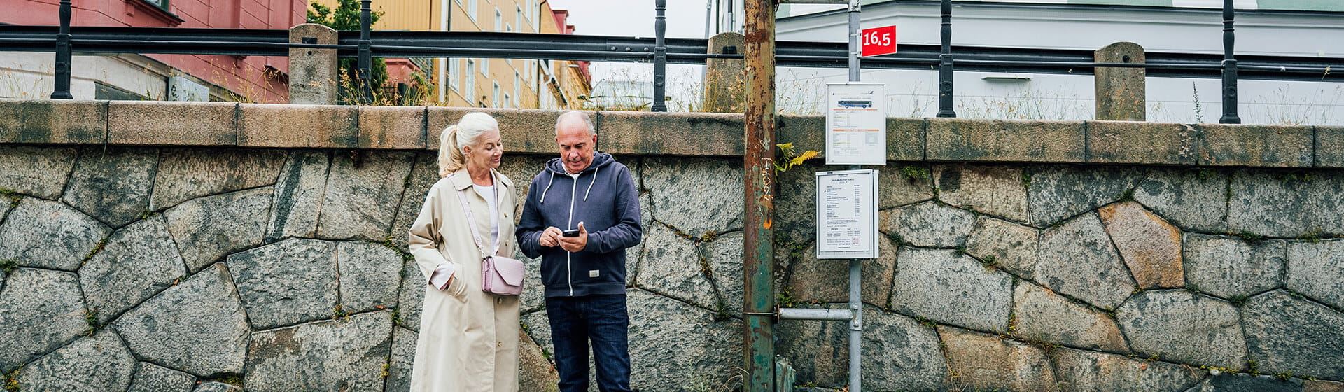 En kvinna och en man står vid en busshållplats. Mannen tittar ner i sin mobiltelefon.