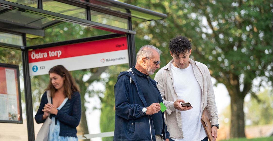 Tre personer står vid en busshållplats.
